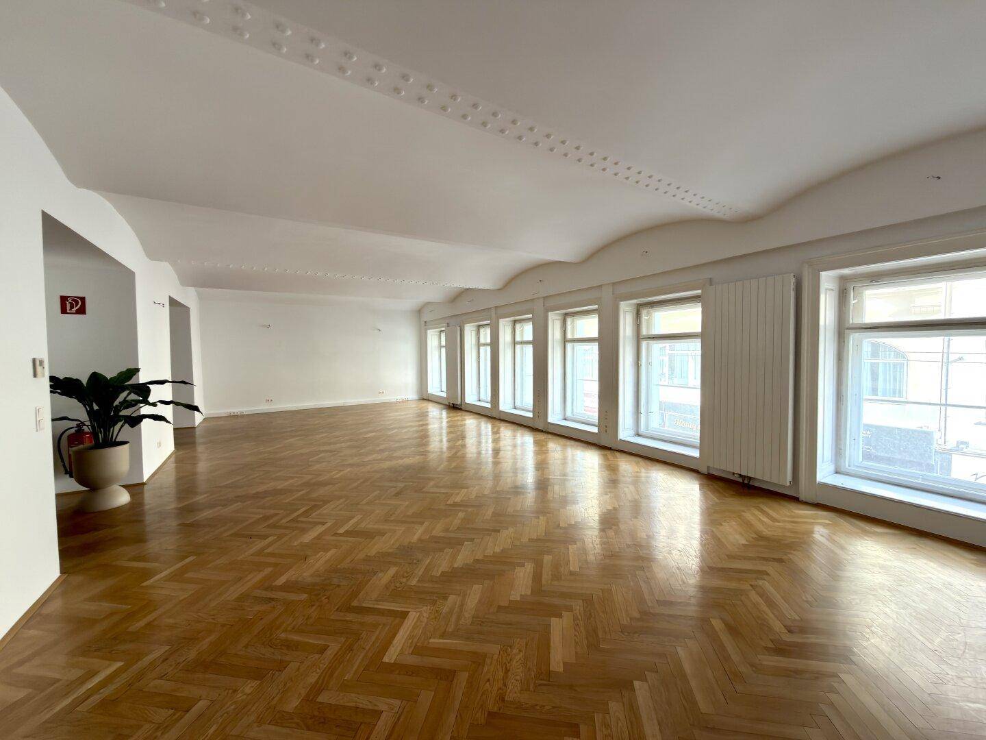 Heller, leerer Büroraum mit Fischgrätparkett, hohen Fenstern und einer eleganten Deckenverzierung, sehr geräumig.