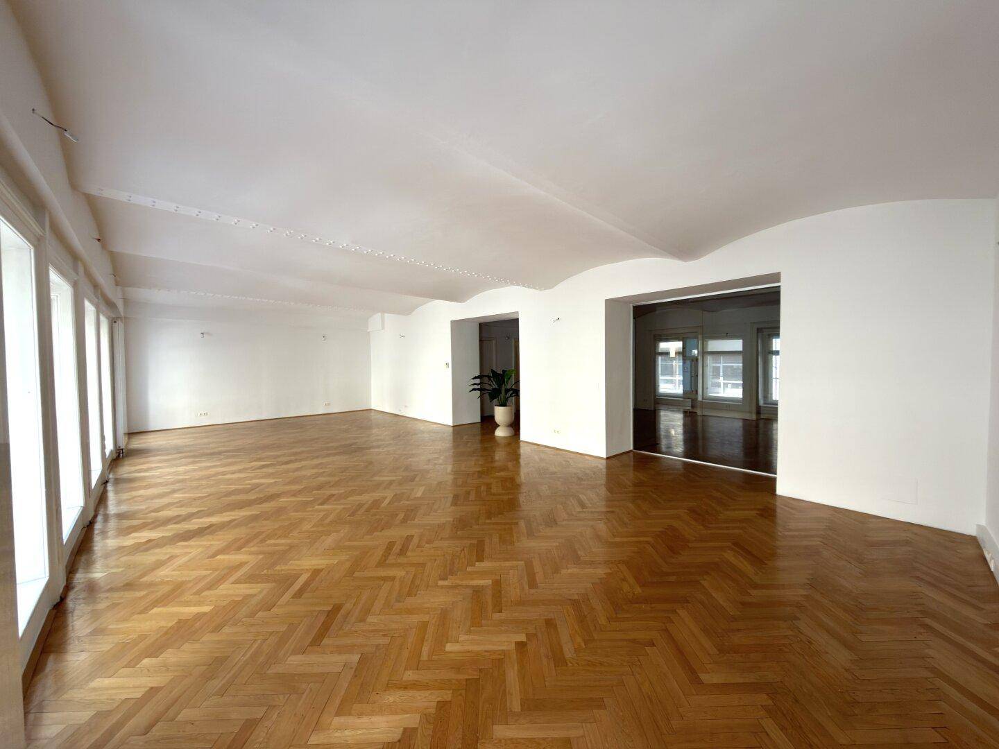 Großer, leerer Büroraum mit glänzendem Fischgrätparkett und einer Reihe von hohen Fenstern, sehr hell.