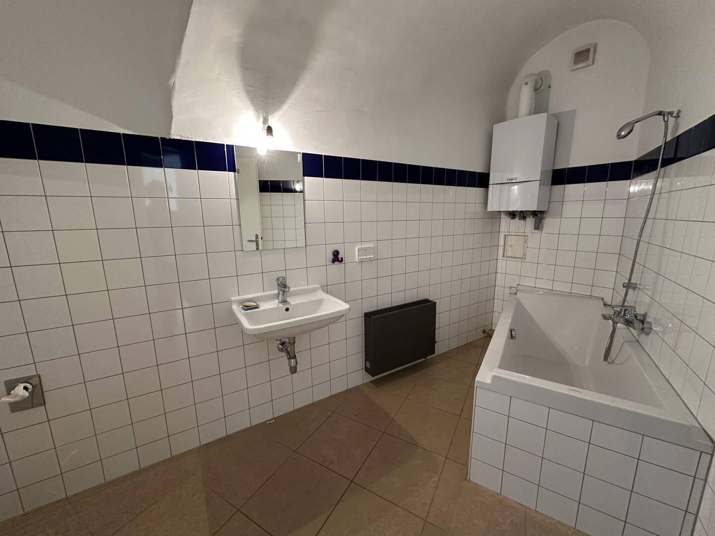 Badezimmer mit weißen Fliesen, blauer Bordüre, Badewanne und Waschbecken unter einem Spiegel.