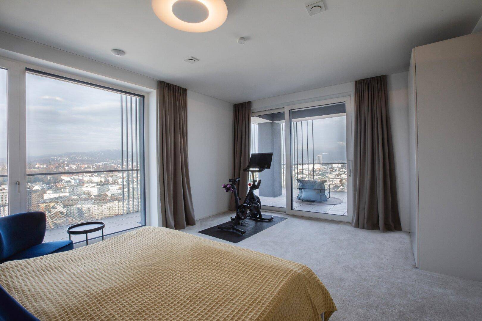Schlafzimmer mit großem Bett, Trainingsgerät und weitem Blick über die Stadt.