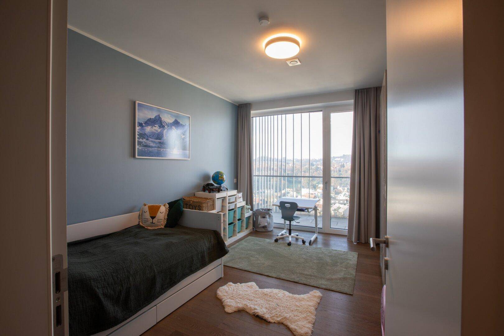 Kinder- oder Gästezimmer mit Bett, Schreibtisch und Blick auf die Stadt durch große Fenster.