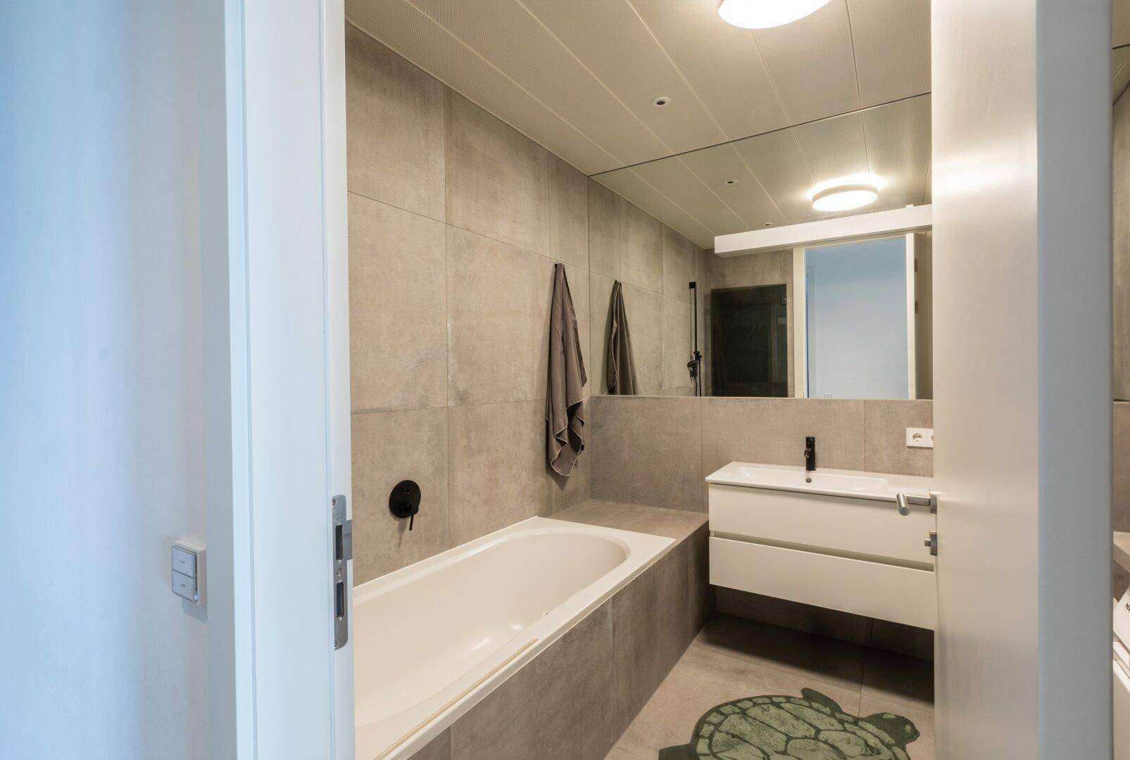 Modernes Badezimmer mit integrierter Badewanne, großem Spiegel und grauen Fliesen.