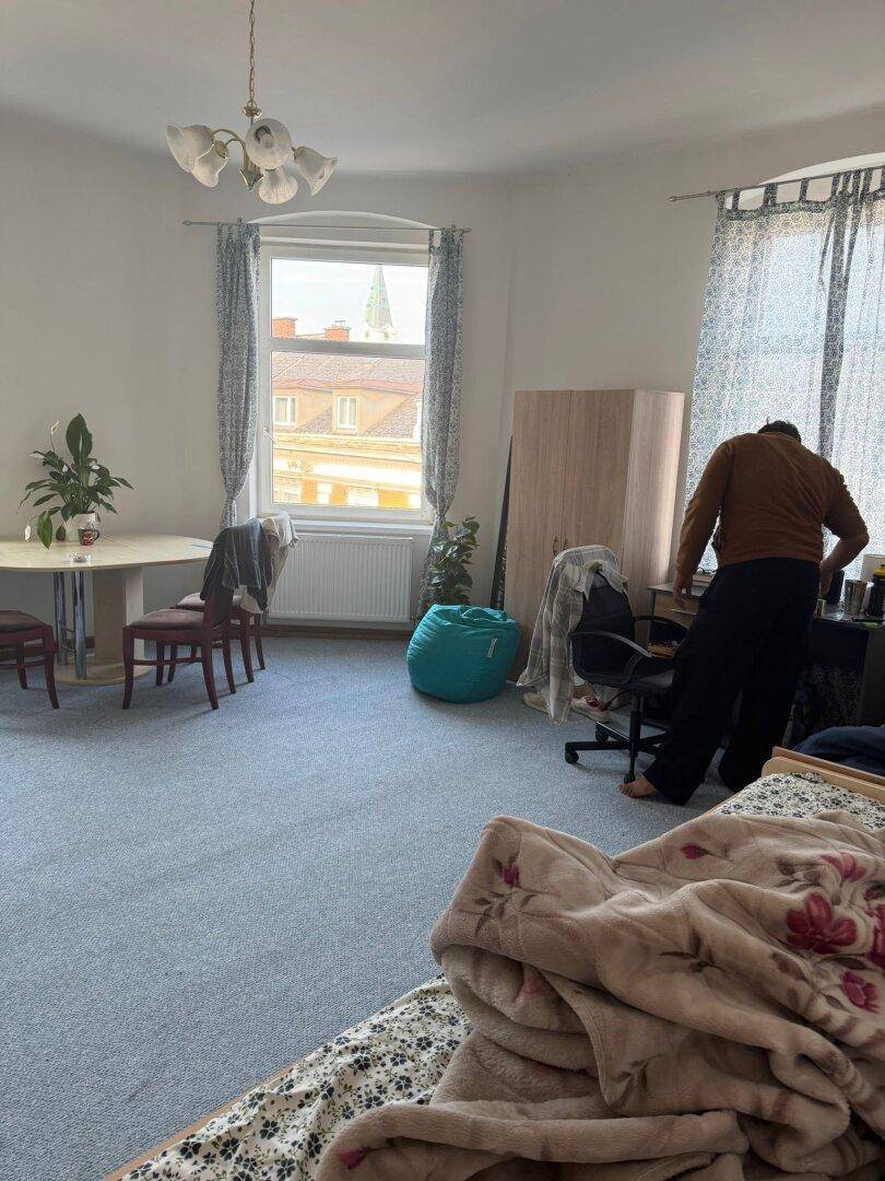 Geräumiges Zimmer mit Teppichboden, einem Esstisch und einem Arbeitsbereich am Fenster.