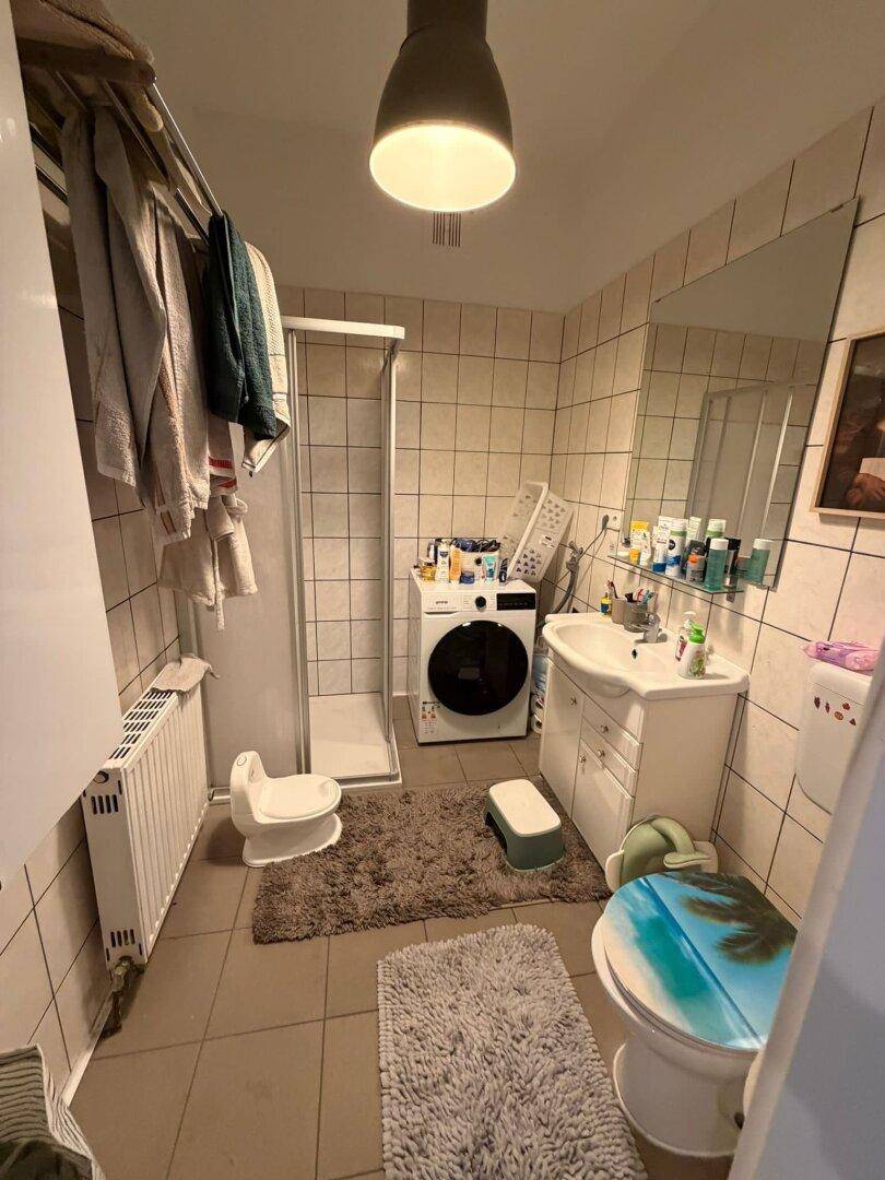 Badezimmer mit Fliesenboden, Dusche, Waschmaschine und einem Waschbecken mit Spiegel.