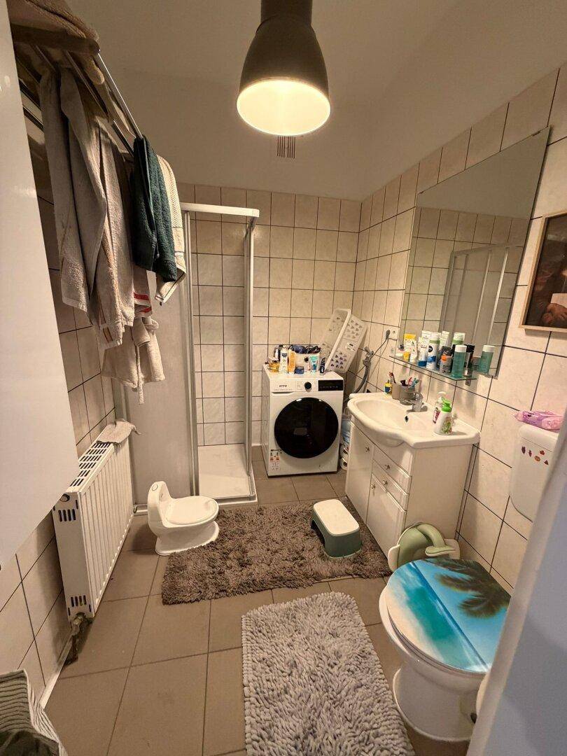 Badezimmer mit Fliesenboden, Dusche, Waschmaschine und einem Waschbecken mit Spiegel.