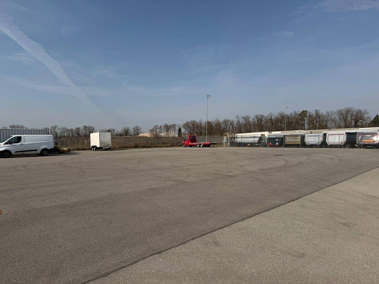 Weitläufiger asphaltierter Außenbereich mit abgestellten Fahrzeugen und Anhängern unter klarem Himmel.