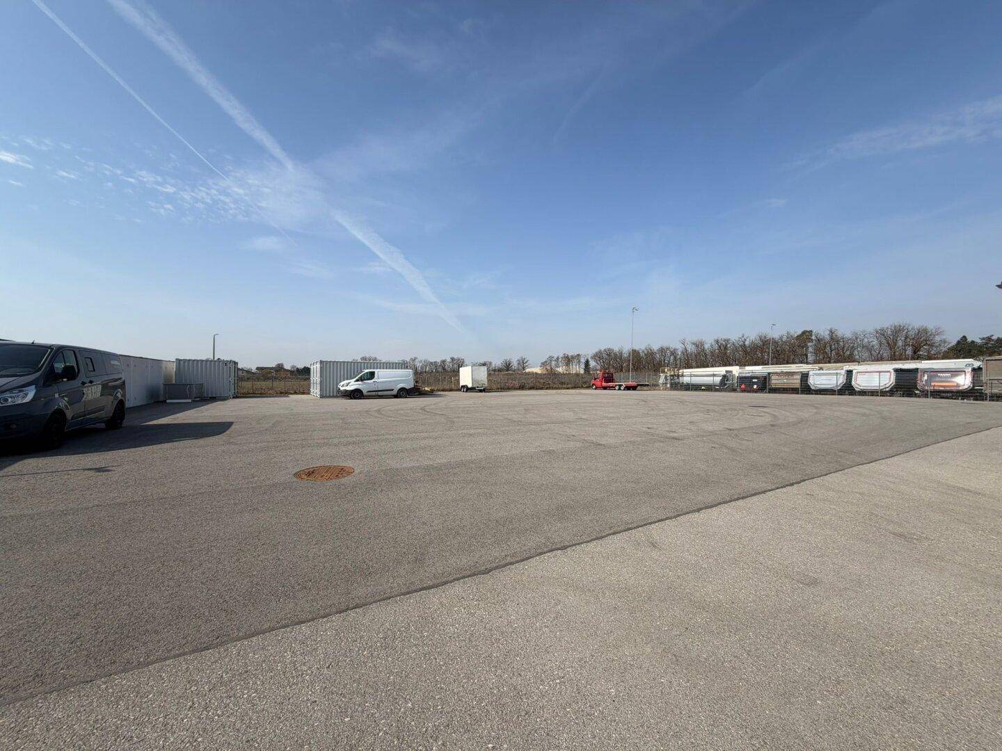 Ausgedehnter asphaltierter Parkbereich mit verschiedenen Fahrzeugen und Containern im Hintergrund.