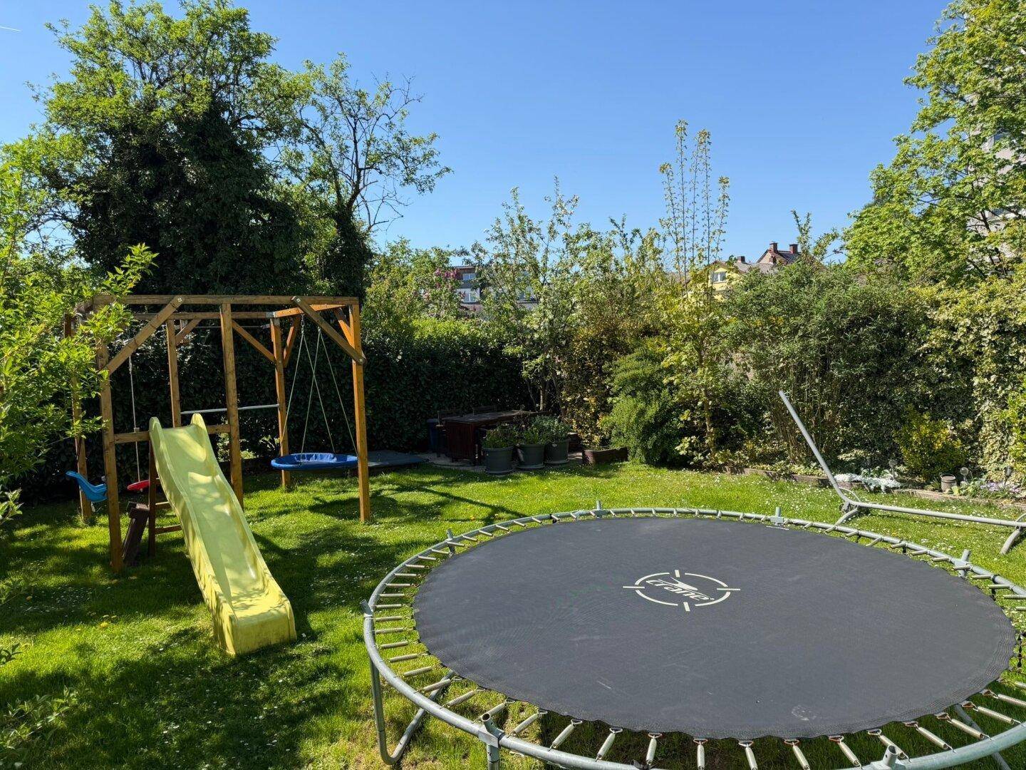 Großer grüner Garten mit Trampolin, Schaukel und Rutsche für Kinder.