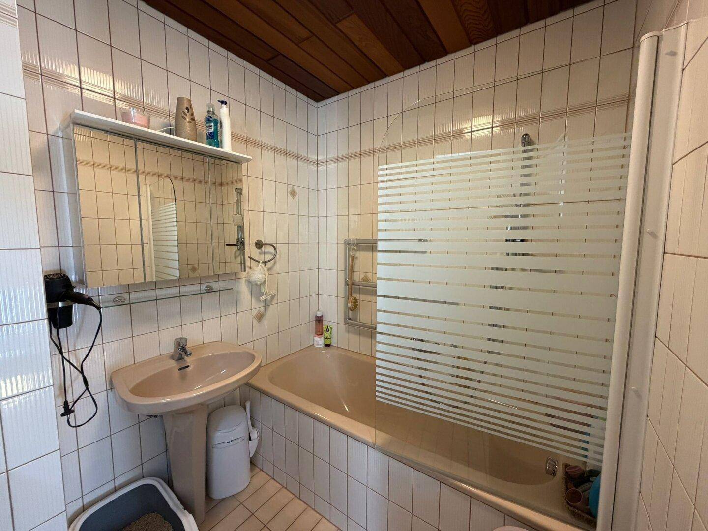 Badezimmer mit Badewanne, Waschbecken und weißen Fliesen im 80er-Jahre-Stil.