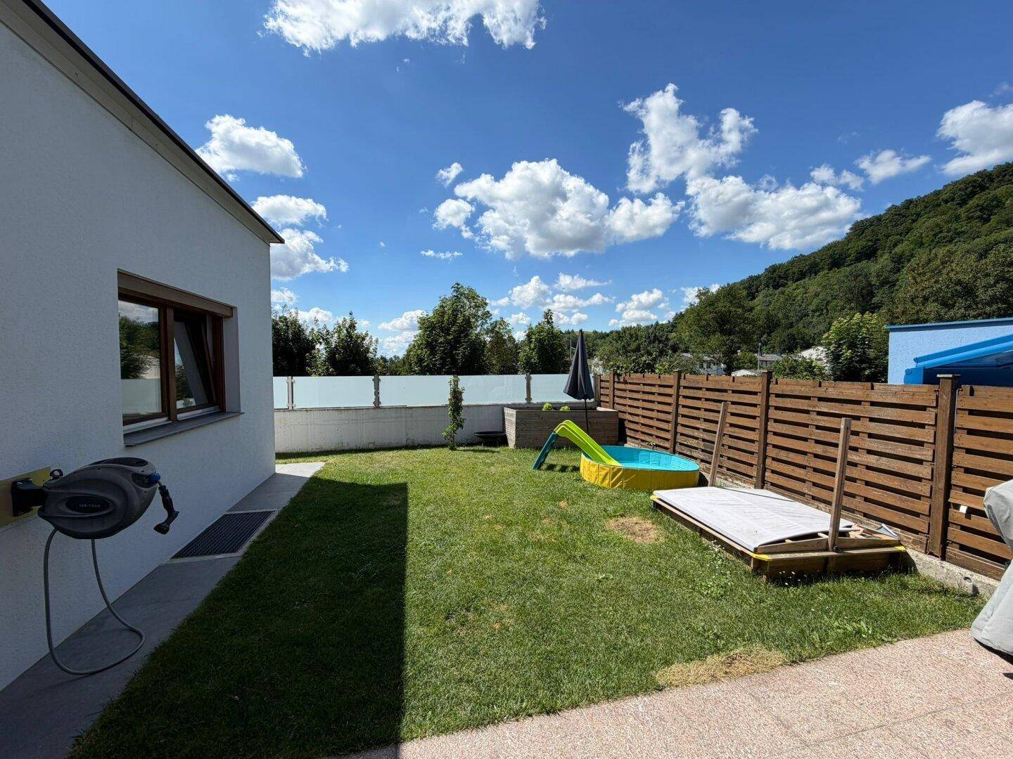 Gartenbereich mit Rasenfläche, kleinem Pool und Holzzaun unter blauem Himmel.