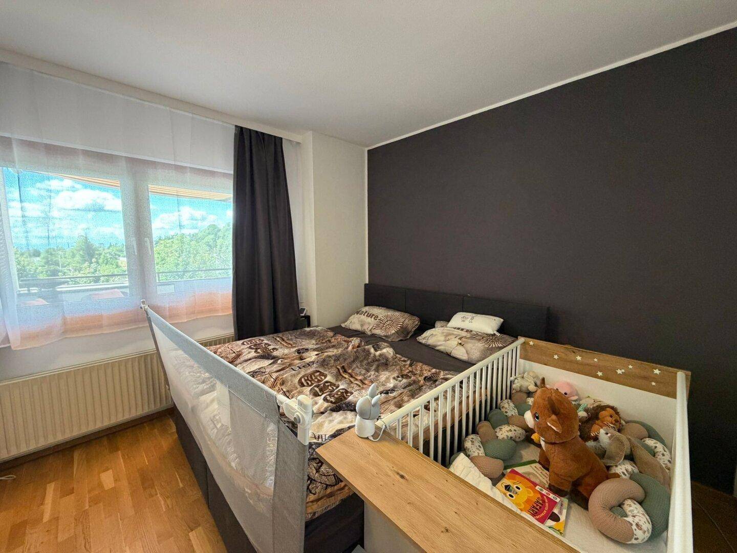 Helles Schlafzimmer mit Doppelbett, Babybett und großem Fenster.