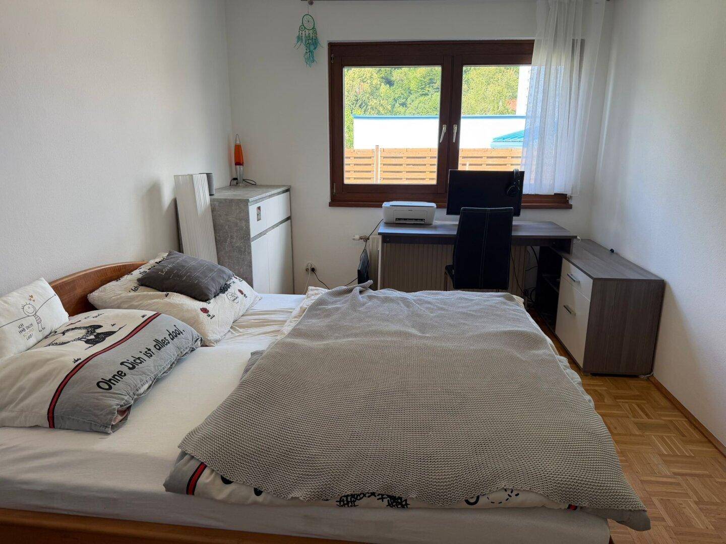 Schlafzimmer mit Bett, Schreibtisch und Fenster mit Blick nach draußen.