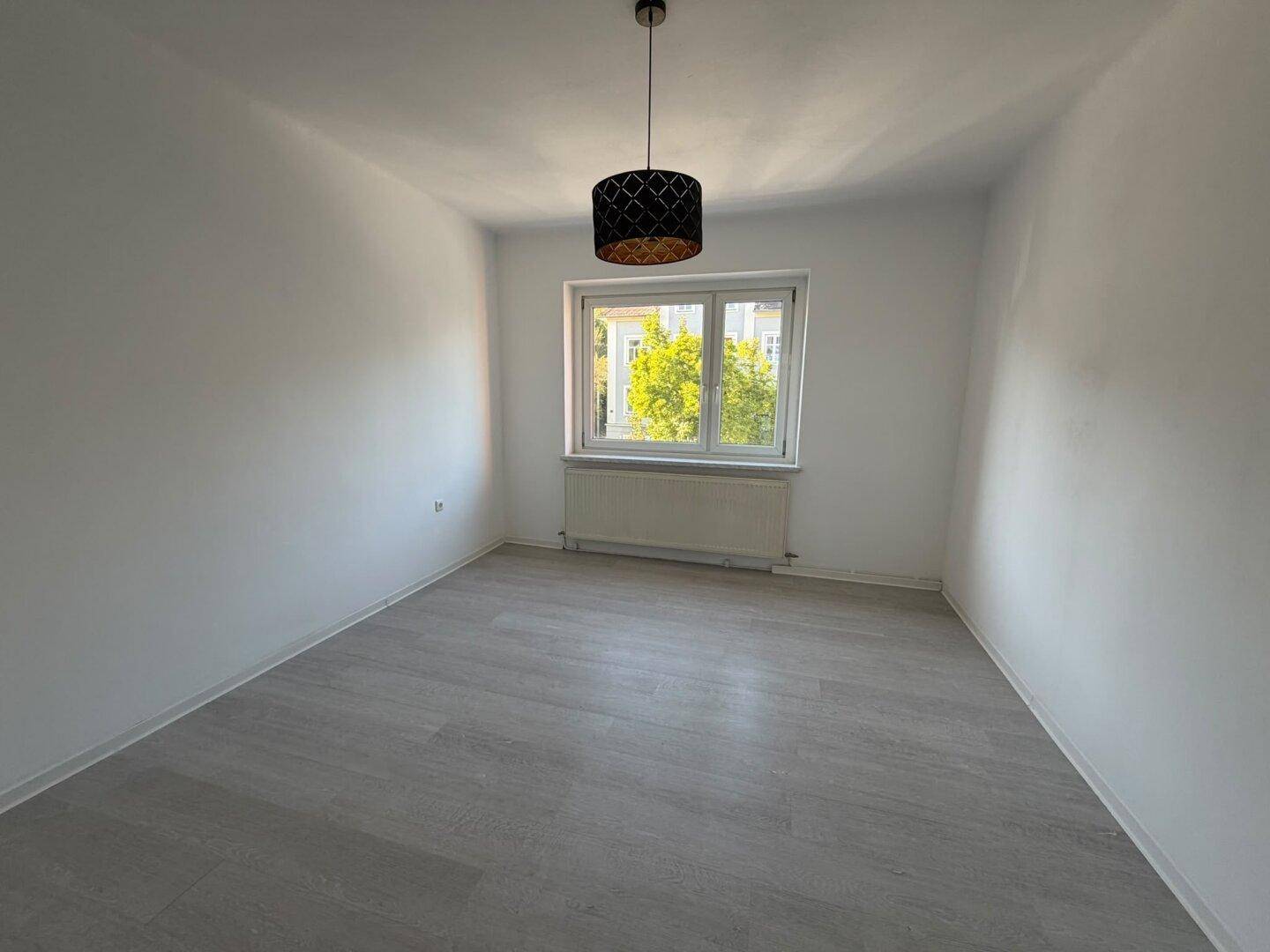 Leeres Schlafzimmer mit hellem Laminatboden, weißen Wänden und einem Fenster mit Blick ins Grüne.