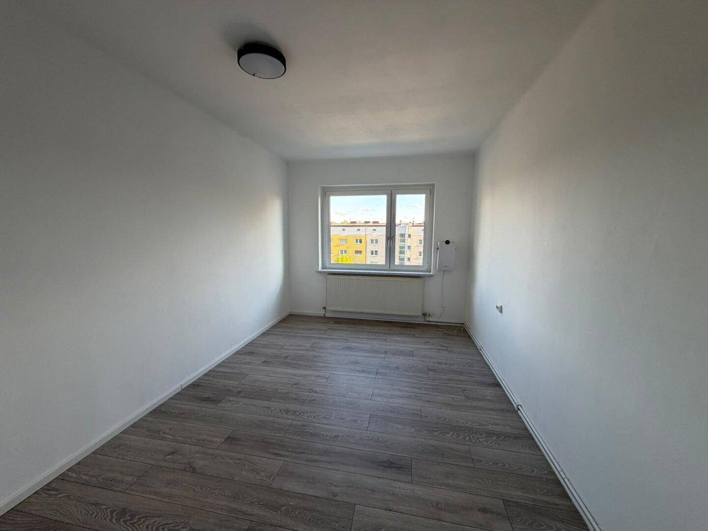 Helles, leeres Zimmer mit dunklem Laminatboden, weißen Wänden und Blick auf die umliegenden Gebäude.
