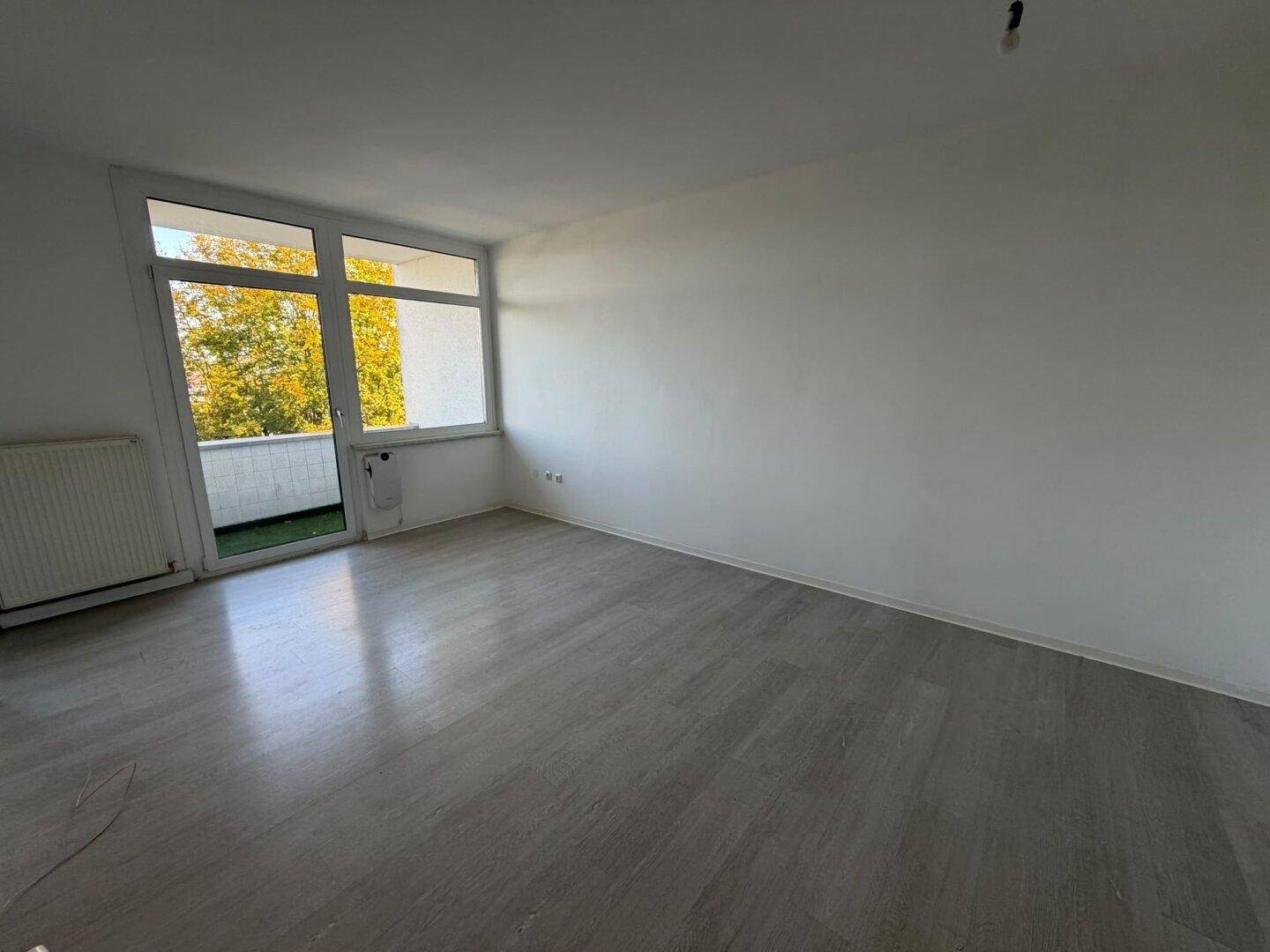 Helles, leeres Zimmer mit Laminatboden, weißen Wänden und Zugang zu einem kleinen Balkon.