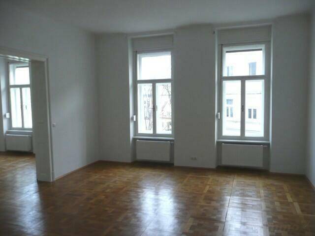 Großes Wohnzimmer mit Parkettboden, zwei Fenstern und offenem Zugang zu einem weiteren Raum.