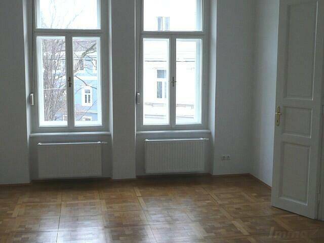 Heller Raum mit Parkettboden und zwei hohen Fenstern, die einen Blick nach draußen bieten.