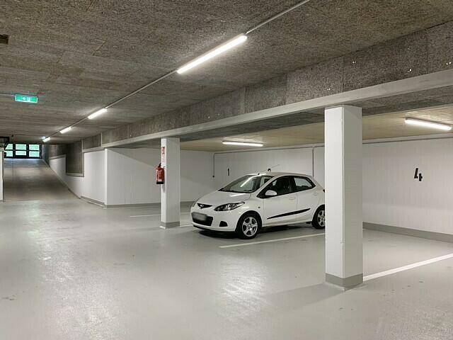Helle Tiefgarage mit mehreren Stellplätzen und einem weißen Auto, die zum Eingang führt.