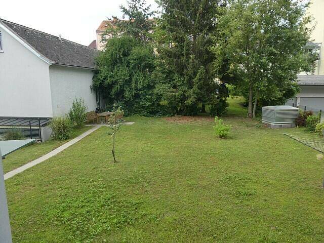 Grüner Gartenbereich mit Rasenfläche, hohen Bäumen und einer kleinen Sitzecke im Hintergrund.
