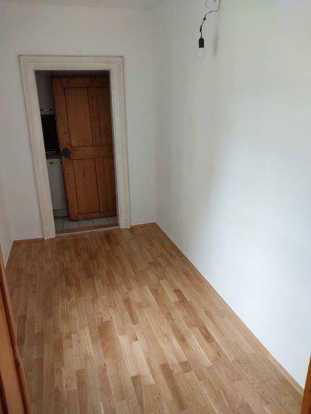 Schmaler Flur oder kleines Zimmer mit hellem Dielenboden und Holztür, führt zu weiteren Räumen.