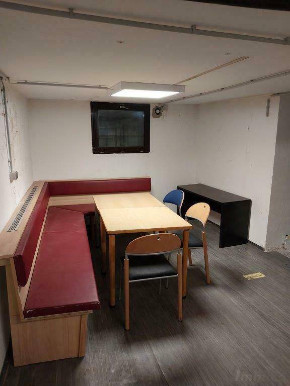 Kellerbüro mit Sitzbank, Tisch und Stühlen, beleuchtet durch eine Deckenleuchte.