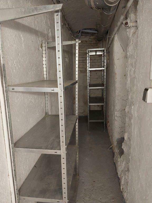 Schmaler Kellergang mit Regalen und sichtbaren Rohren an der Decke.