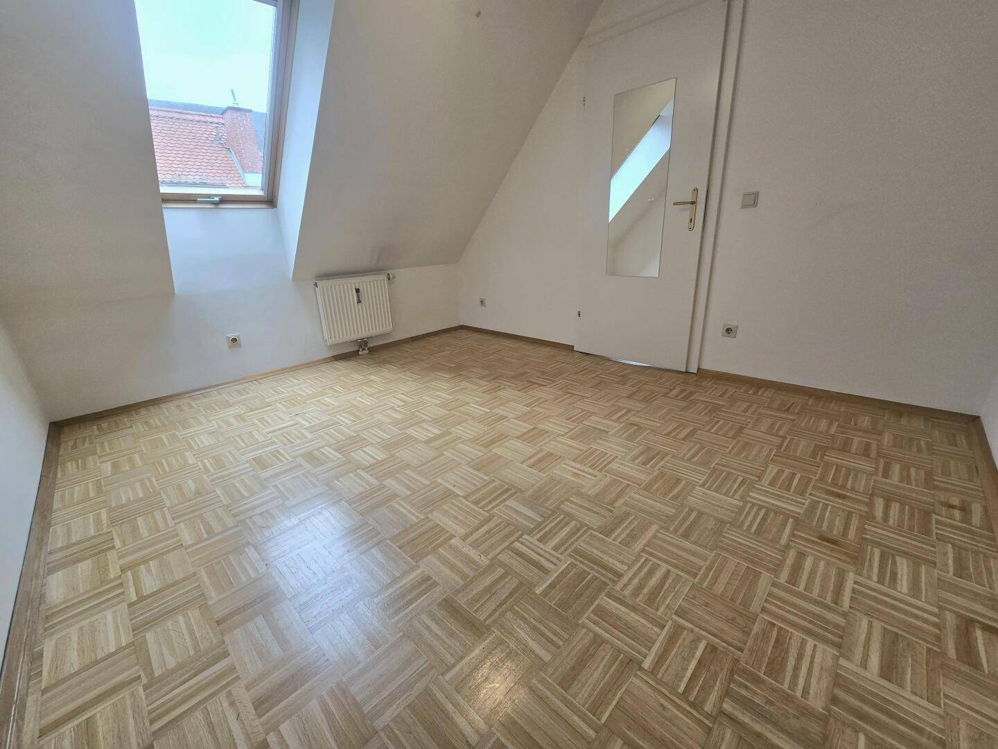 Helles Zimmer mit Parkettboden, Dachschräge und Tür mit Spiegel.