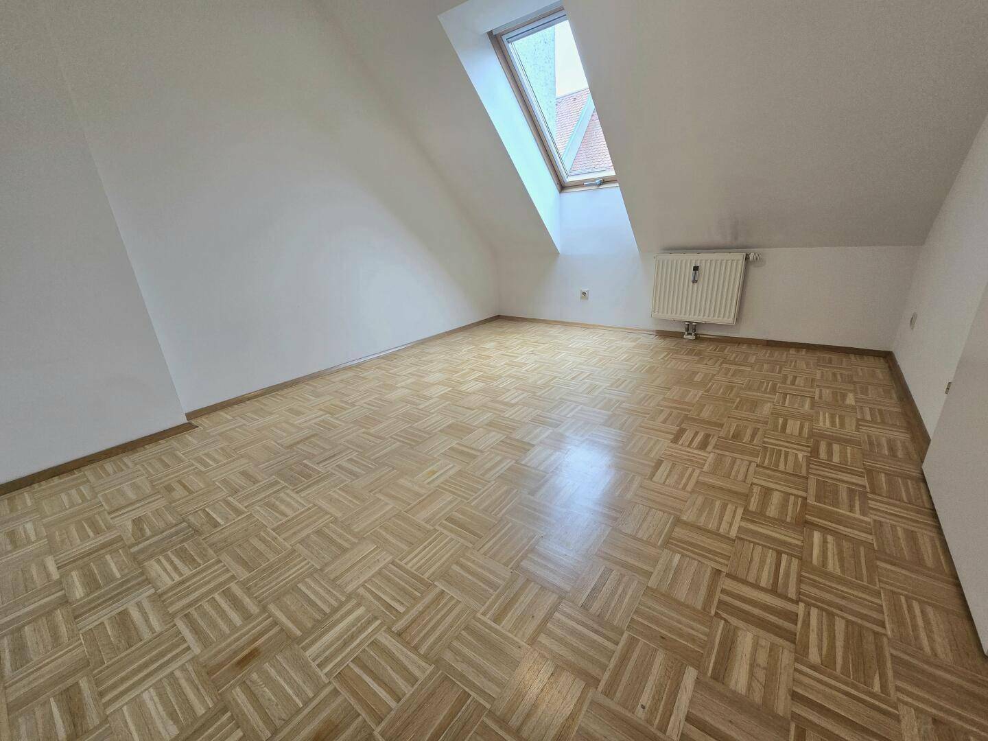 Leeres Schlafzimmer mit Parkettboden, Dachschräge und einem Dachfenster.