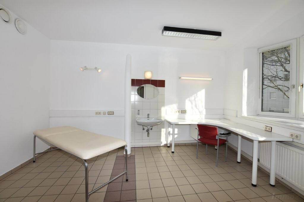 Funktionaler Praxisraum mit Behandlungsliege, Schreibtisch und großem Fenster für viel Tageslicht.