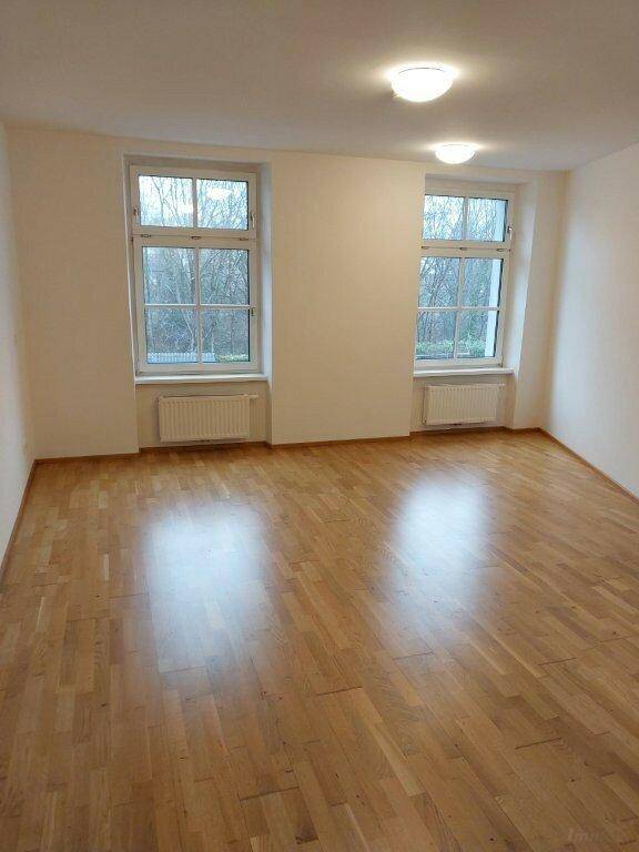 Heller Wohnraum mit Parkettboden, zwei großen Fenstern und weiß gestrichenen Wänden.