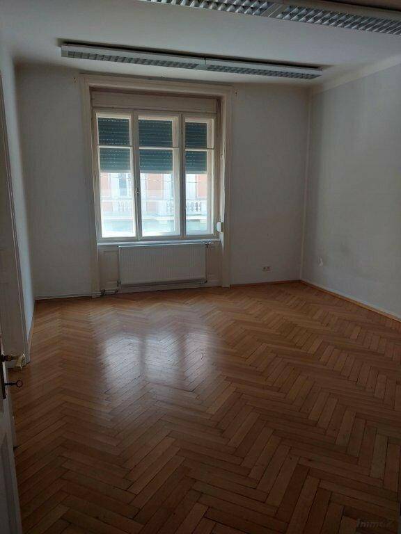 Geräumiges Zimmer mit glänzendem Fischgrätparkett und einem großen Fenster mit Rollladen.
