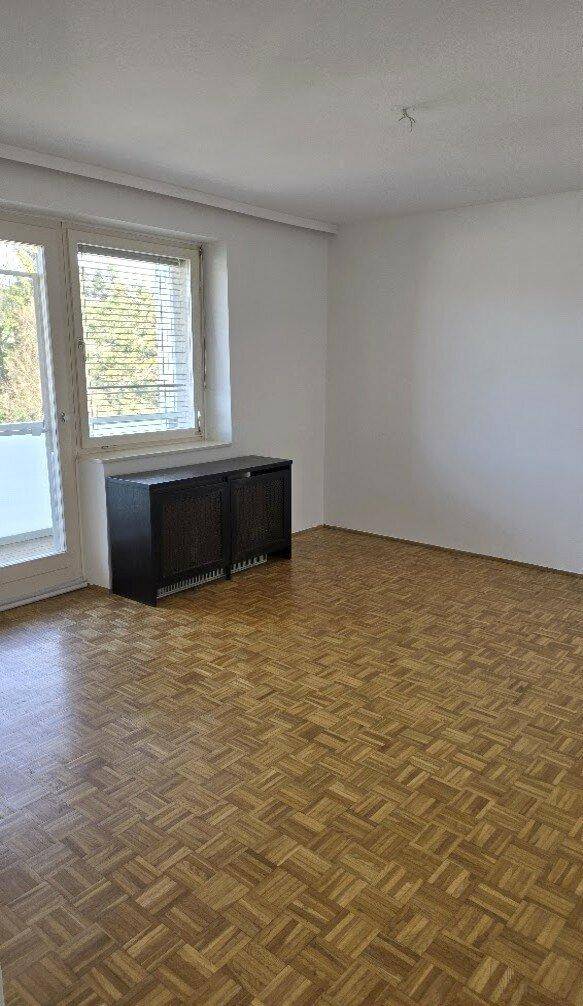 Leeres Zimmer mit Parkettboden und großem Fenster, das viel Tageslicht hereinlässt.