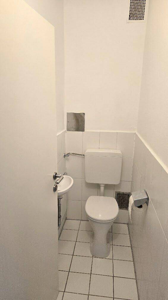 Separate Toilette mit kleinem Waschbecken und weißen Wand- und Bodenfliesen.