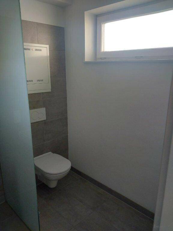 Separates WC mit wandhängender Toilette, Fenster und grauen Wandfliesen.