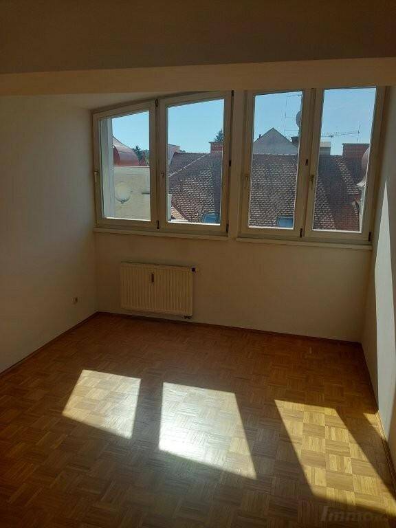Sonniger Raum mit Parkettboden und Fenstern, die einen Blick auf die umliegenden Dächer bieten.