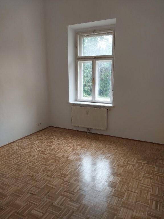 Helles Zimmer mit Blockparkettboden und einem großen Fenster, das den Blick ins Grüne freigibt.