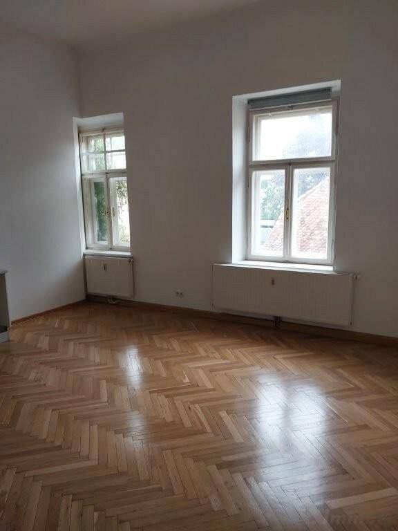 Heller Wohnraum mit klassischem Fischgrätparkett, hohen Decken und zwei großen Fenstern.