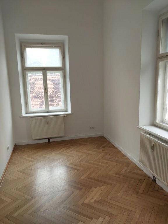 Leeres Zimmer mit klassischem Fischgrätparkett und zwei Fenstern, die eine helle Atmosphäre schaffen.