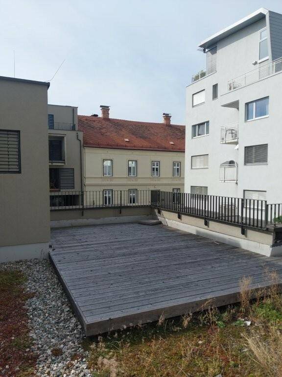 Eine Holzterrasse im Freien mit Blick auf umliegende moderne Gebäude und einen begrünten Bereich.