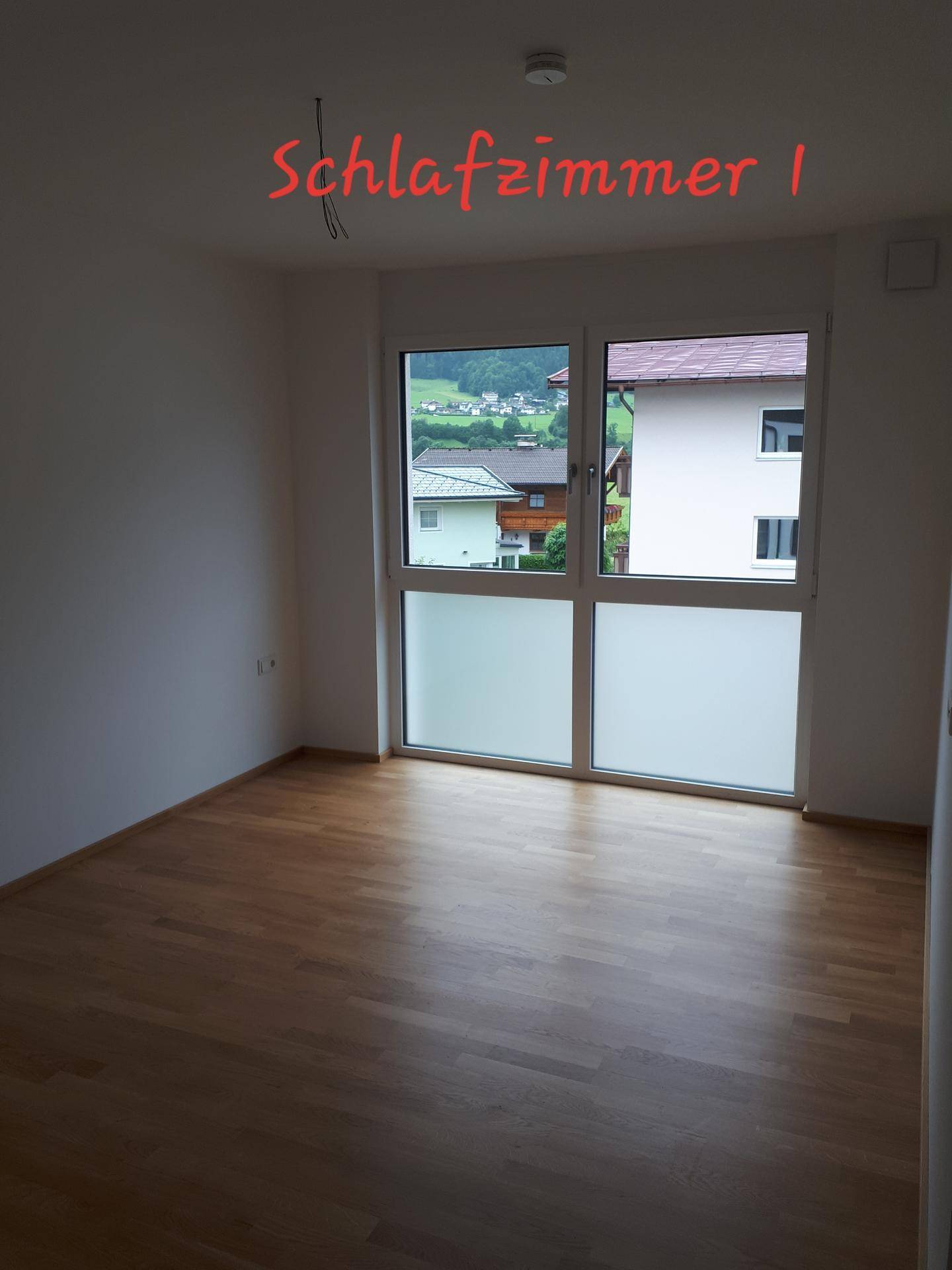 Helles Schlafzimmer mit Holzparkettboden und großem Fenster mit Blick auf die umliegenden Häuser und Natur.