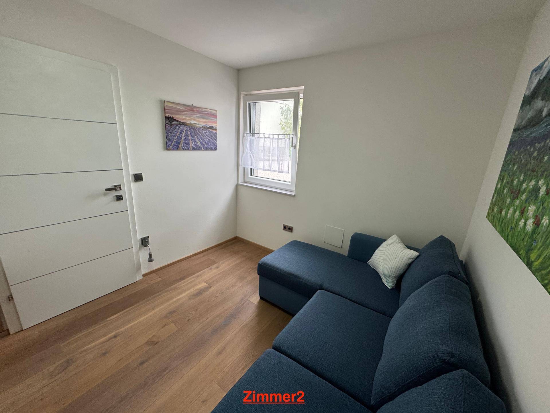 Helles Zimmer mit blauem Sofa, Holzboden und weiß gestrichenen Wänden.