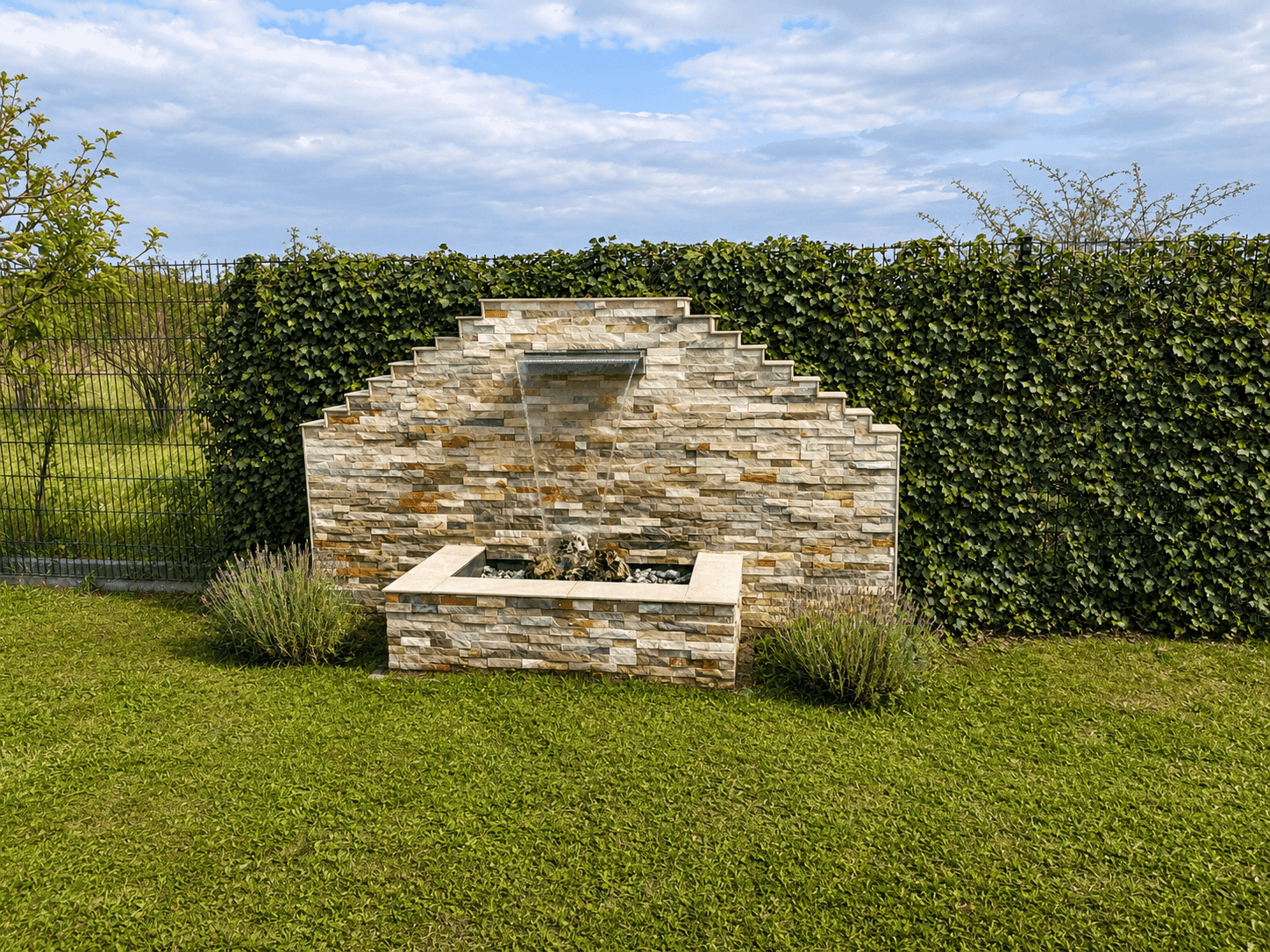 Dekorativer Steinbrunnen im Garten, umgeben von grünen Pflanzen und einer dichten Efeuwand.