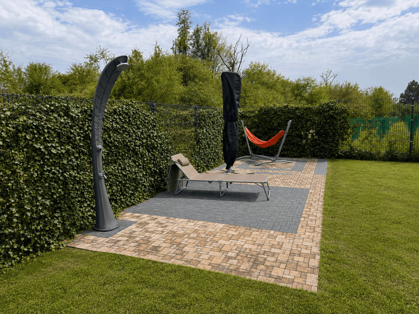 Gemütlicher Außenbereich im Garten mit Außendusche, Sonnenliege und Hängematte auf gepflasterter Fläche.