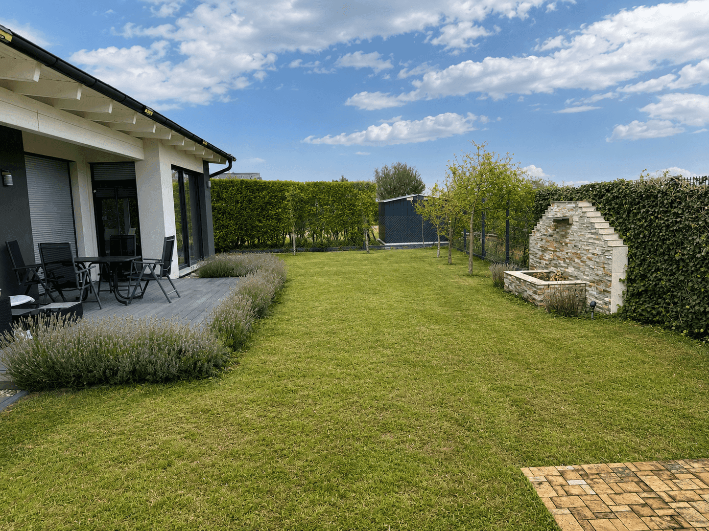 Gepflegter Garten mit Rasenfläche, Terrasse und dekorativem Steinbrunnen, umgeben von Hecken.