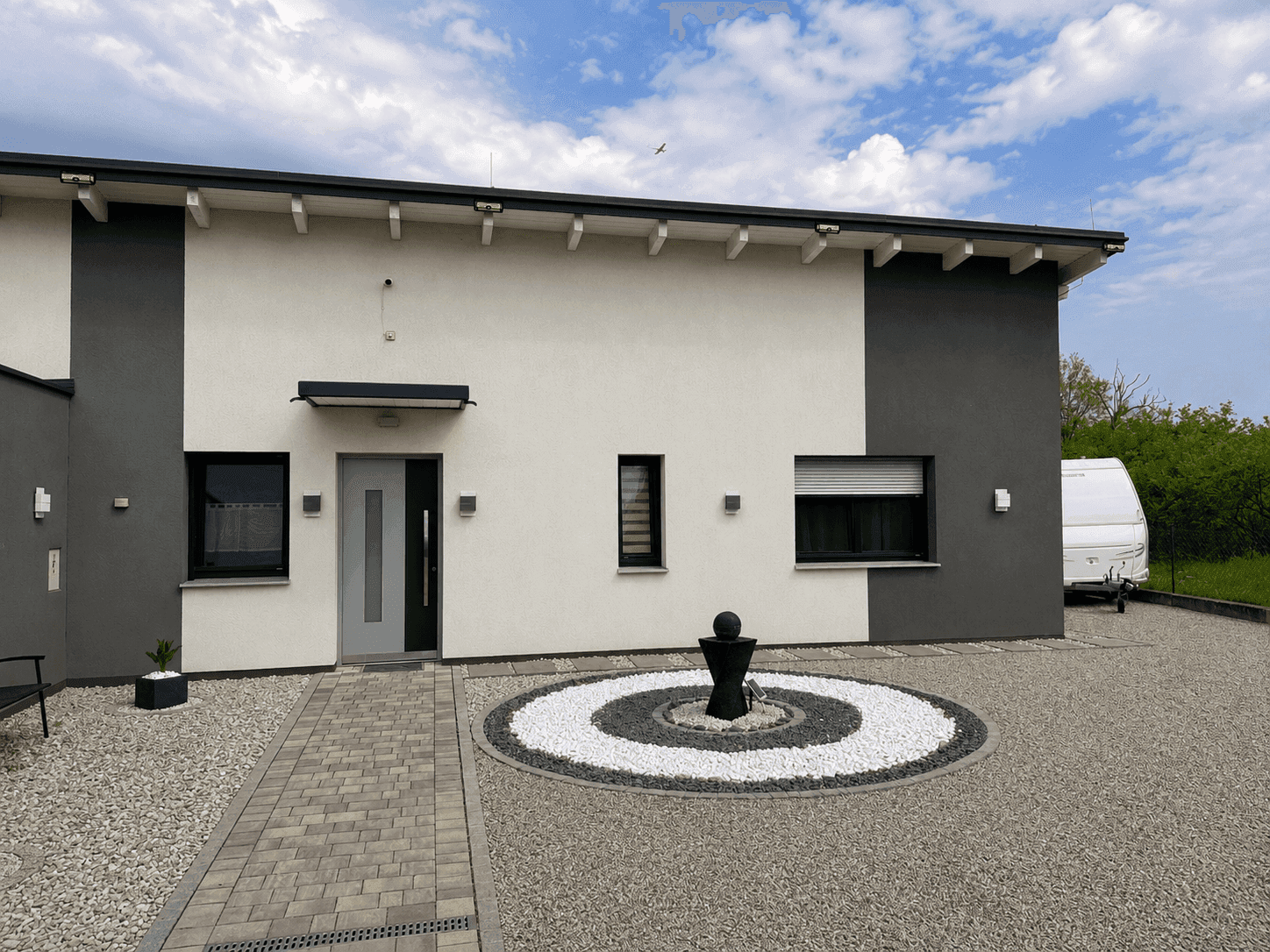 Eingangsbereich des modernen Hauses mit grauer Tür, Zierbrunnen und gepflegter Kiesfläche.