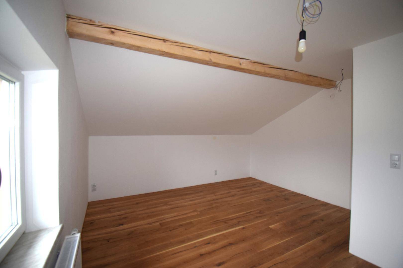 Leerer Raum im Dachgeschoss mit Holzbalken, schräger Decke und Holzdielenboden, hell durch ein Fenster.