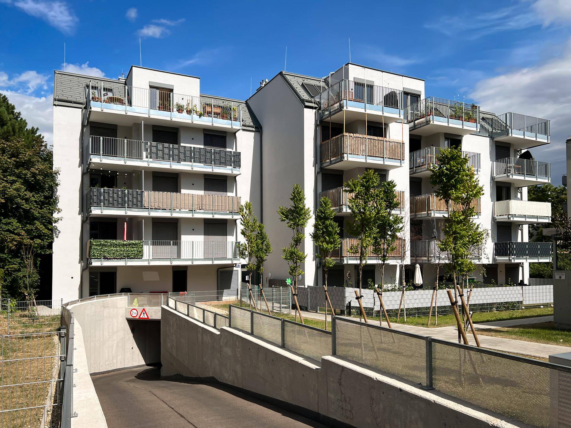Modernes Mehrfamilienhaus mit Balkonen und Zufahrt zur Tiefgarage unter blauem Himmel.