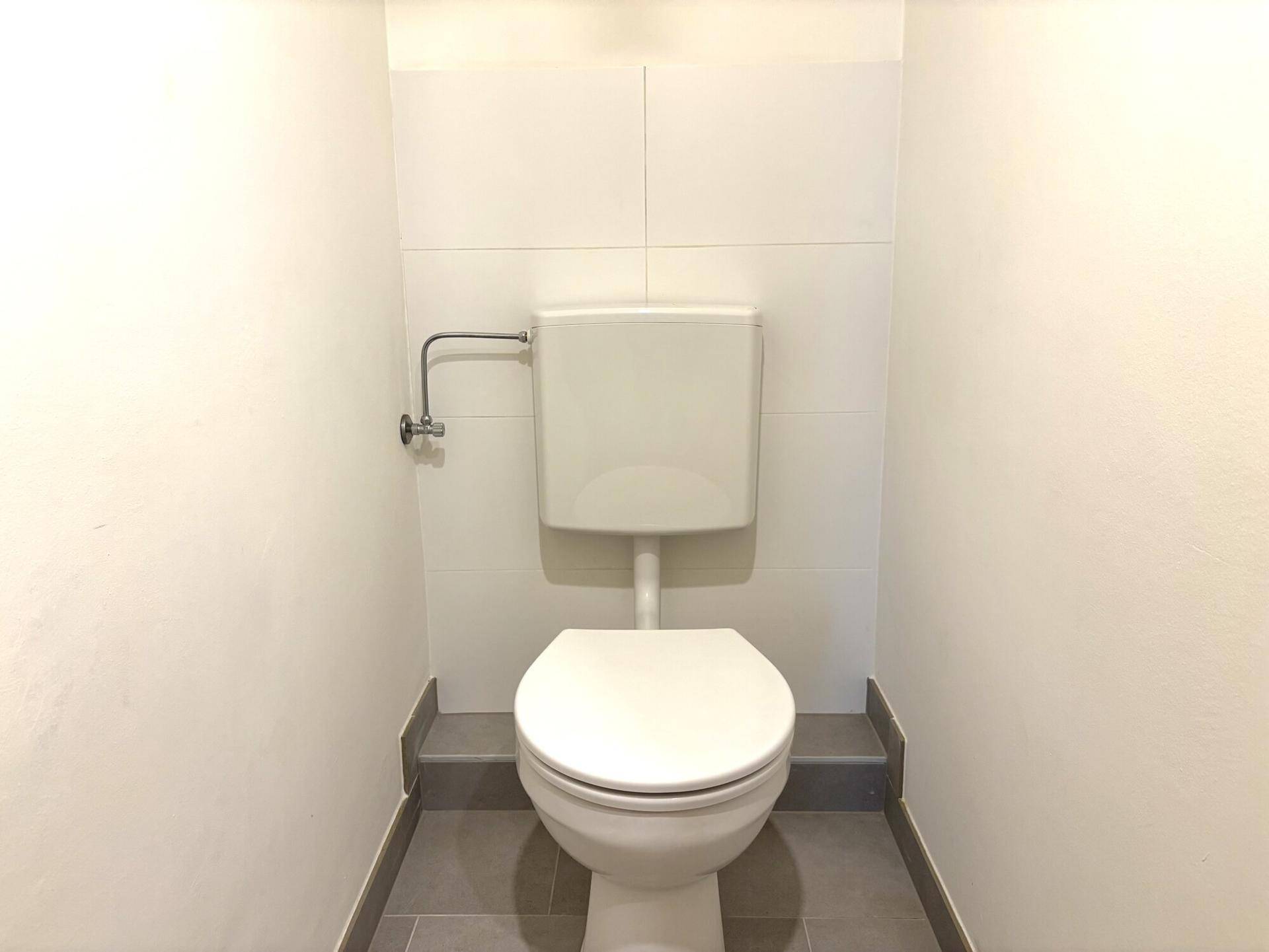 Separates WC mit Toilette und weißen Fliesen an den Wänden sowie grauen Bodenfliesen.