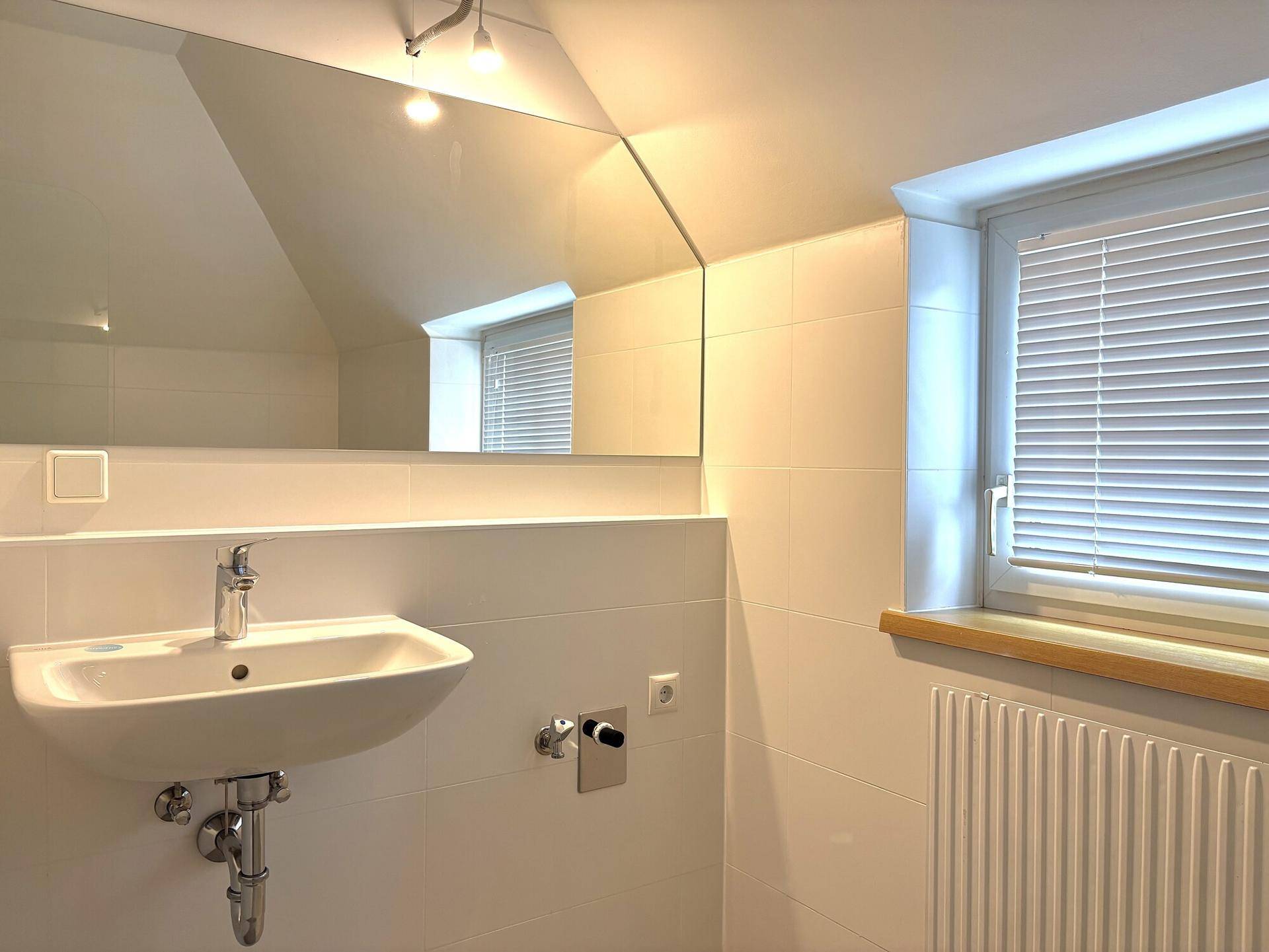 Helles Badezimmer mit Waschbecken, großem Spiegel und Fenster mit Jalousie unter der Dachschräge.