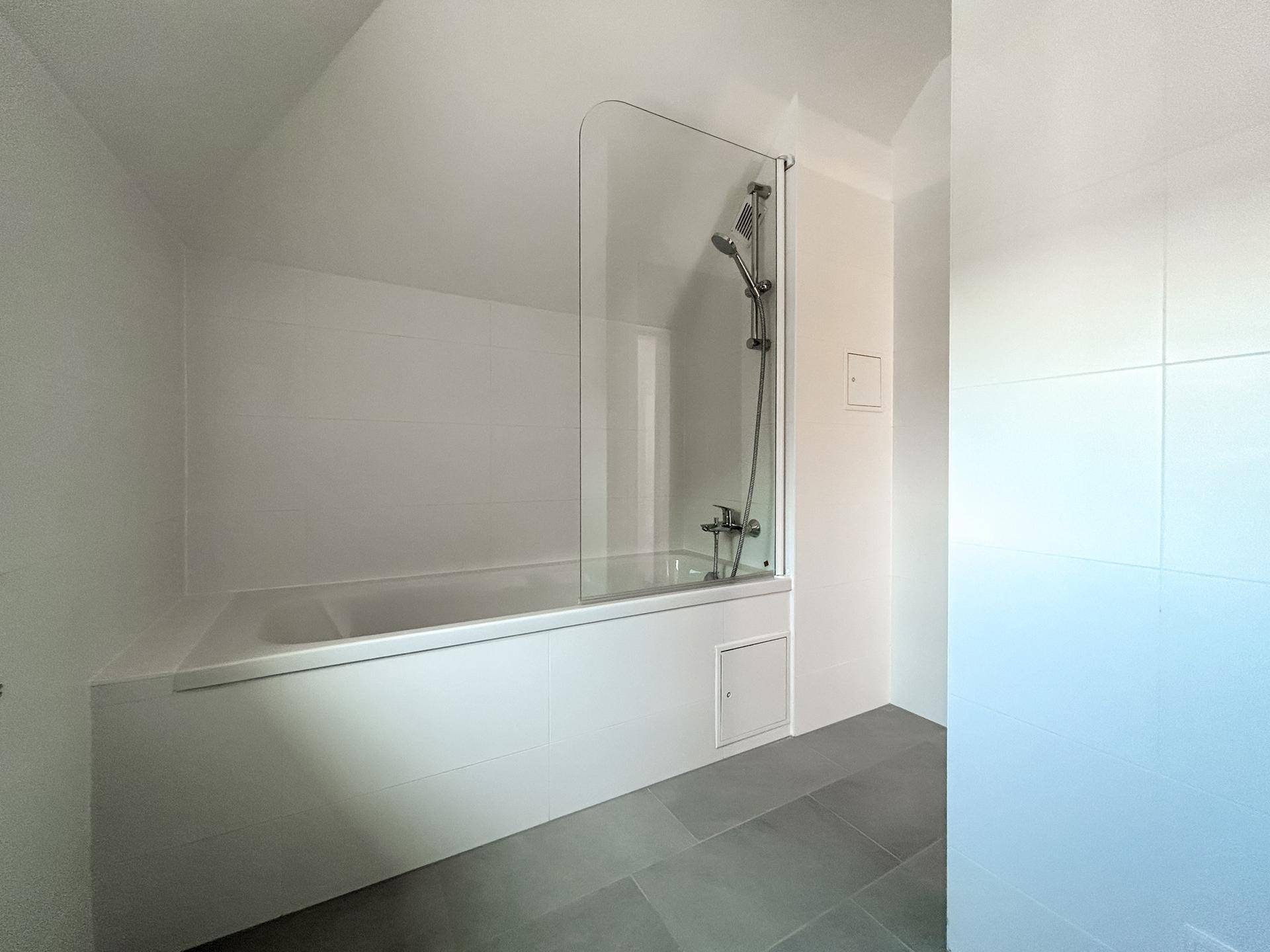 Modernes Badezimmer mit Badewanne, Duschwand und weißen Wandfliesen sowie grauen Bodenfliesen.